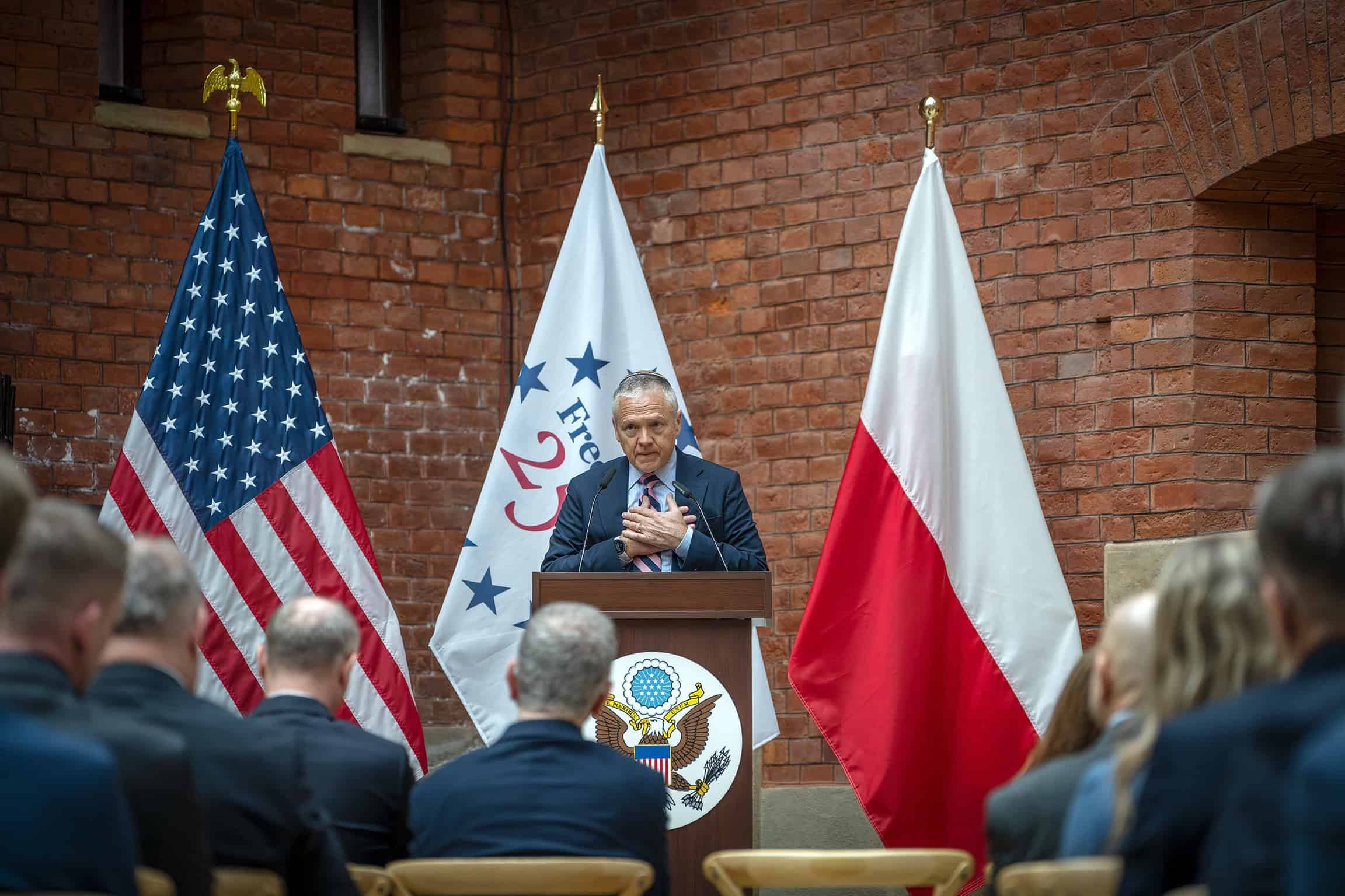 Ambasador USA przemawia podczas wydarzenia w Komitecie Kopca Kościuszki, z flagami Polski i USA w tl.
